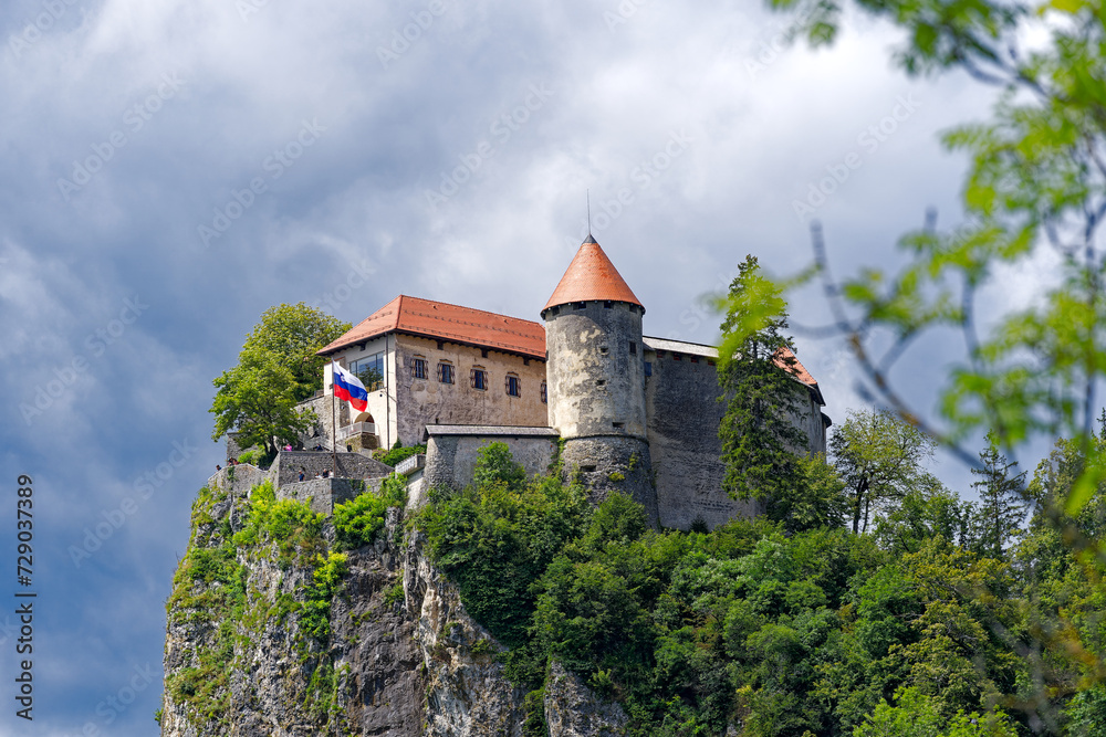 Scenic view of castle on top of rock with Slovenian flag waving at lakeshore of Slovenian Lake Bled on a cloudy summer day. Photo taken August 8th, 2023, Bled, Slovenia.