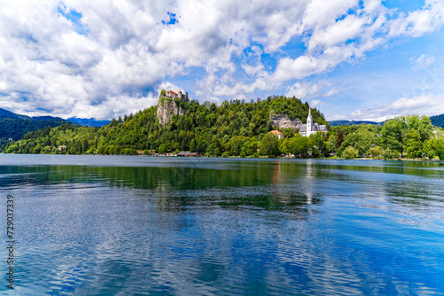 Scenic view of Lake Bled with beautiful reflections and castle on a rock and woodland in the background on a blue cloudy summer day. Photo taken August 8th, 2023, Bled, Slovenia.