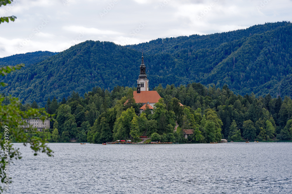 Lake Bled with church on an island and woodland in the background on a cloudy summer day. Photo taken August 8th, 2023, Bled, Slovenia.