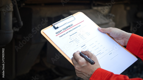  Action of a mechanic engineer is checking on heavy machine checklist form to verify the quality of maintenance service, with train locomotive part as blurred background. Industrial working scene.