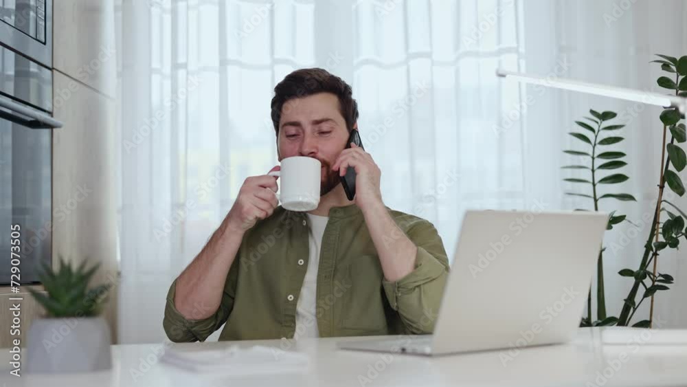 Handsome freelancer beginning working day with cup of coffee while talking on smartphone at desk with modern laptop. Contented man in casual wear enjoying business conversation and hot drink at home. Handsome freelancer beginning working day with cup of coffee while talking on smartphone at desk with modern laptop. Contented man in casual wear enjoying business conversation and hot drink at home.