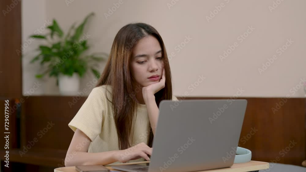 Asian young adult with bored expression, laptop in use, feeling uninspired, sitting at home. Woman with blue headphones, staring at laptop screen, lack of enthusiasm, wooden table, plant aside Asian young adult with bored expression, laptop in use, feeling uninspired, sitting at home. Woman with blue headphones, staring at laptop screen, lack of enthusiasm, wooden table, plant aside