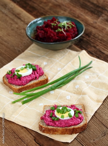 Beetroot mixed with goat cream cheese spreaded on whole grain bread. Decorated with slice of egg and green onion. Red beetroot salad and herbs at back.
