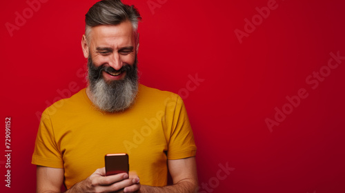 bearded older man with a top knot hairstyle, laughing and looking at his phone, wearing a yellow t-shirt against a solid red background