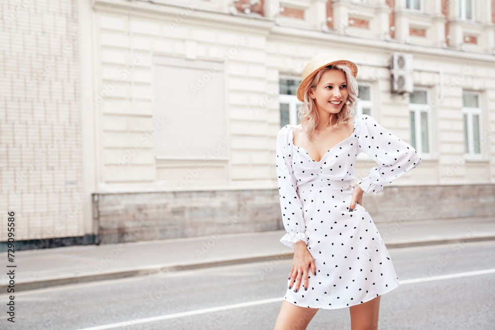Young beautiful smiling blond woman in trendy summer white dress. Sexy carefree woman posing in the street at sunset. Positive model outdoors at sunny day. Cheerful and happy. In hat