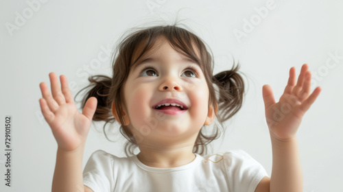 joyful toddler with pigtails is raising her hands in excitement and looking up, set against a plain light background