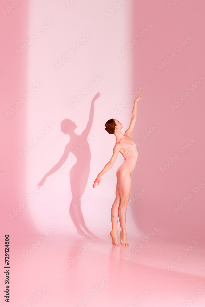 Pink Elegance. Woman in sporty swimsuit dancing elegantly against pastel studio background. Shadow captures her grace. Concept of ballet, beauty, elegance, movement, shadowplay.