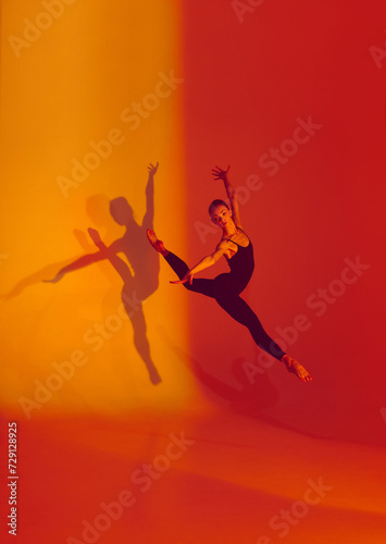 Fototapeta Naklejka Na Ścianę i Meble -  Elegant Motion in Neon. Graceful dancer poses in black sports overalls, barefoot against gradient red-orange backdrop, her shadow adding depth to her movements. Concept of grace, athleticism, motion.