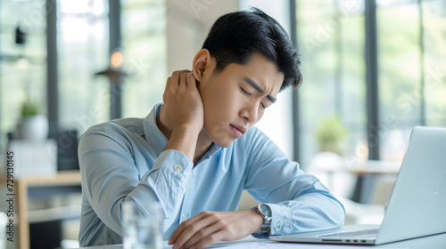 Young man is experiencing discomfort or neck pain while working on a laptop in a bright office setting. photo