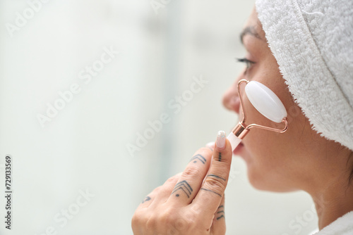 Young woman with soft white towel on head having procedure of facial massage with jade stone roller after morning shower in bathroom