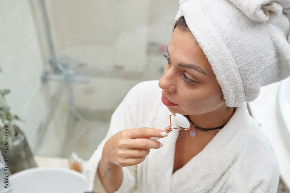 Girl with soft white towel on head and jade stone roller in hand having procedure of facial massage in front of mirror in bathroom Girl with soft white towel on head and jade stone roller in hand having procedure of facial massage in front of mirror in bathroom