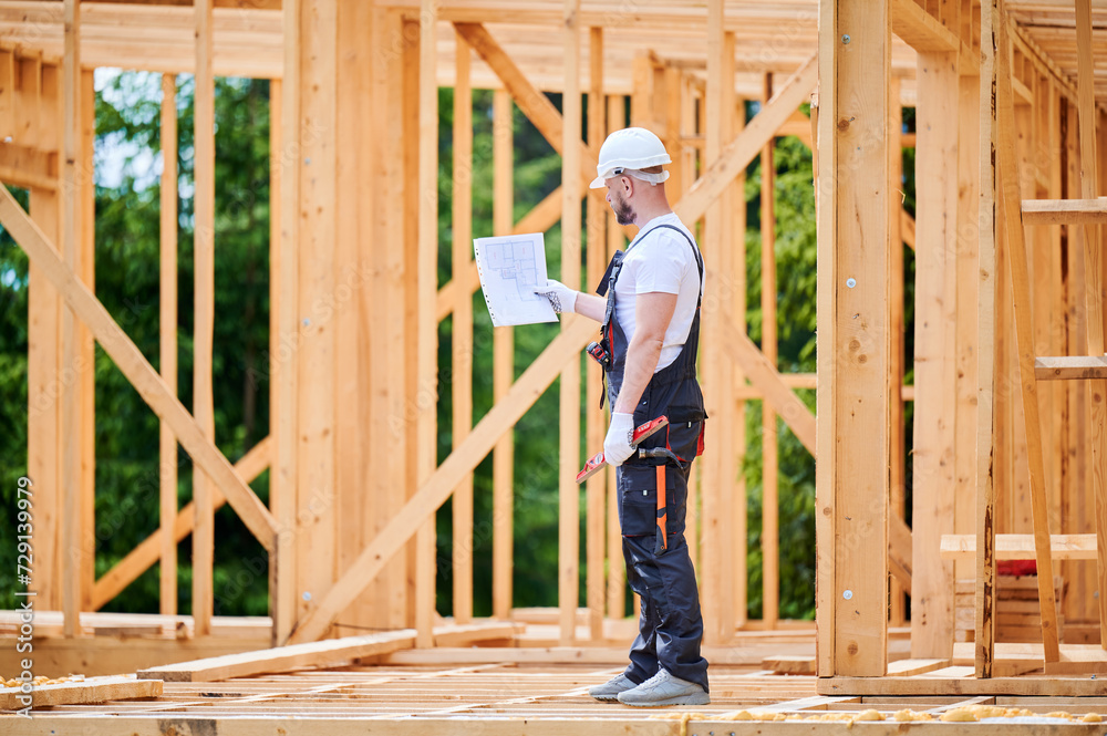 Construction worker building two-story wooden frame house. Man with beard, wearing jumpsuit and hard hat, reviewing the building plan. Intention to implement modern ecological construction practices. Construction worker building two-story wooden frame house. Man with beard, wearing jumpsuit and hard hat, reviewing the building plan. Intention to implement modern ecological construction practices.