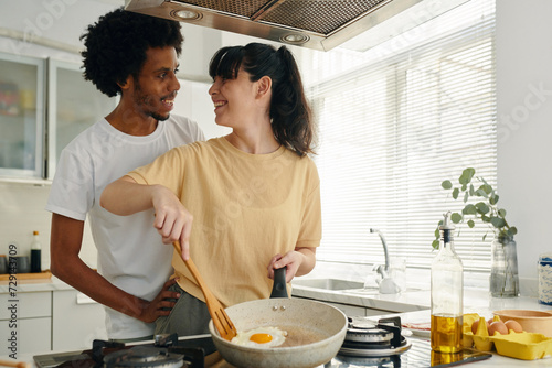Pretty young woman with frying pan cooking scrambled eggs for breakfast while standing by electric stove and looking at her husband photo