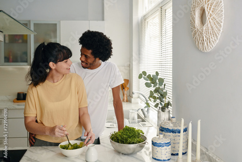 Happy young intercultural husband and wife in t-shirts looking at each other while woman mixing salad ingredients in bowl