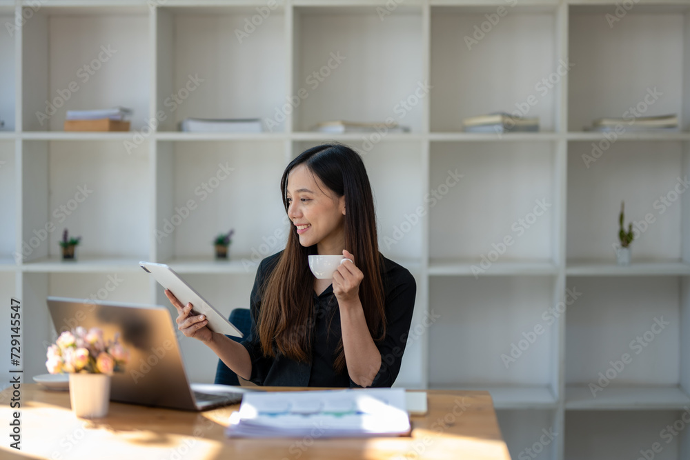 Smiling businesswoman enjoying a cup of coffee while browsing on a digital tablet at her office workspace.. Smiling businesswoman enjoying a cup of coffee while browsing on a digital tablet at her office workspace..