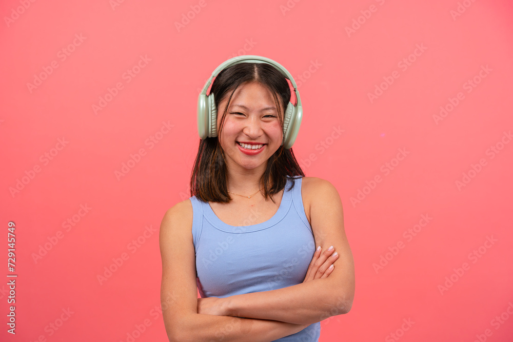 Happy Chinese woman with headphones on pink background, with arms crossed looking at camera