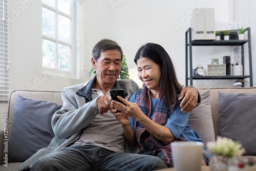 Happy elderly asian couple using smartphone sit on sofa doing ecommerce shopping online on website at home