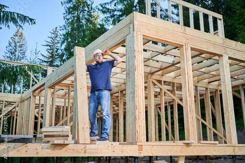 Carpenter constructing wooden two-story frame house near the forest. Bearded man with glasses holding beam, donning protective helmet. Concept of environmentally friendly modern construction.