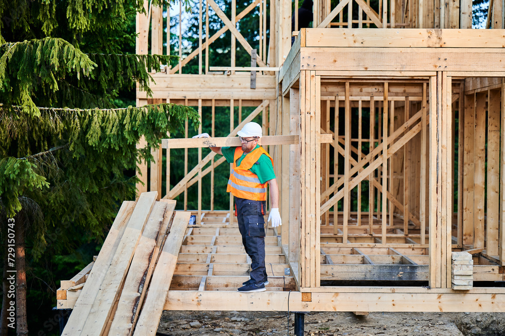 Carpenter building wooden frame house near the forest. Man holds large truss on his shoulder, dressed in work clothes and helmet. Concept of contemporary and sustainable construction. Carpenter building wooden frame house near the forest. Man holds large truss on his shoulder, dressed in work clothes and helmet. Concept of contemporary and sustainable construction.