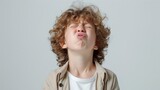 A young child with curly hair making a playful face with closed eyes and puckered lips wearing a light-colored jacket over a white shirt against a neutral background.
