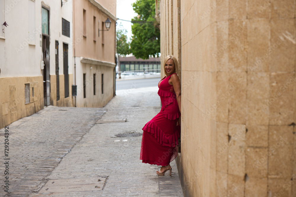 Beautiful blonde mature woman in an elegant dress leaning against the wall of a building on a narrow street in a town in Andalusia, Spain.