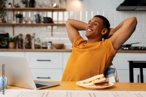 Smiling young African American man stretching while sitting in modern kitchen at home tasty sandwich