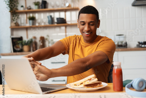 Portrait of smiling young African American man using laptop, stretching, snack break, tasty sandwich