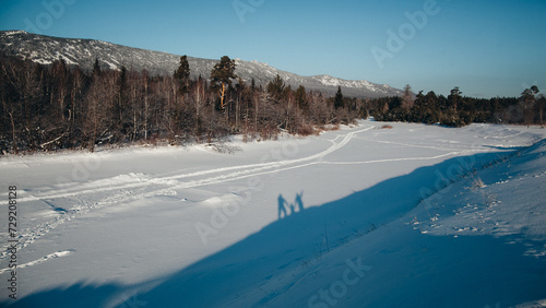 Couple walking on frozen lake, snow-covered trees, and distant mountains under clear blue sky