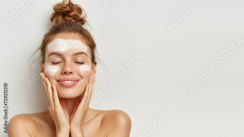 Positive young beautiful woman in white top with closed eyes applying moisturizing cream on face. High-quality crop photo of skincare and cosmetics concept with copy space for text.