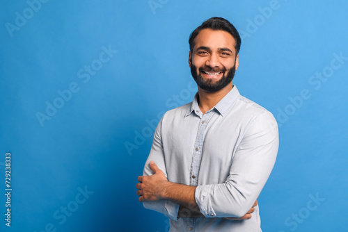 In front of a bright blue background, the Indian businessman stands with arms crossed, his casual light blue shirt suggesting a relaxed yet confident business approach