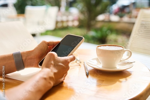 Young woman sitting in coffee shop at wooden table, drinking coffee and using smartphone.On table is laptop. Girl browsing internet, chatting, blogging. Female holding phone and looking on his screen.