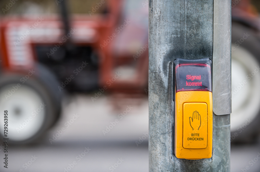 Gelber Druckschalter an Ampelanlage für Fußgänger mit Leuchtschrift Signal kommt vor vorbeifahrendem Traktor
