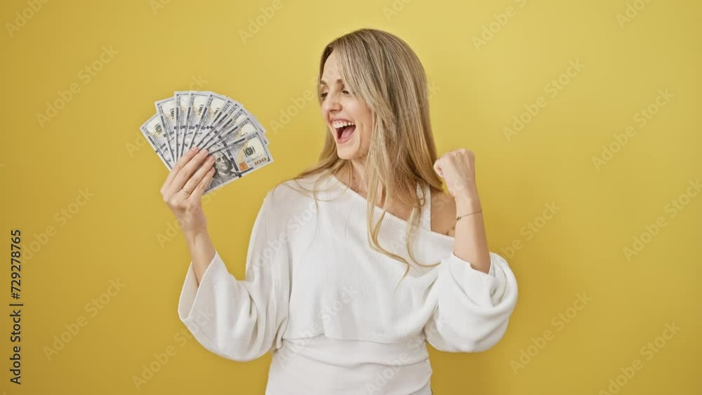 Cheering young blonde woman holding dollars in victory, excited over major success. raised arms in proud triumph against isolated yellow background. Cheering young blonde woman holding dollars in victory, excited over major success. raised arms in proud triumph against isolated yellow background.