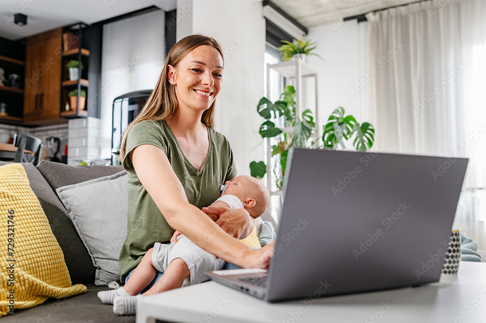 Young new mom holding cute baby in her arms at home while working on a computer Young new mom holding cute baby in her arms at home while working on a computer