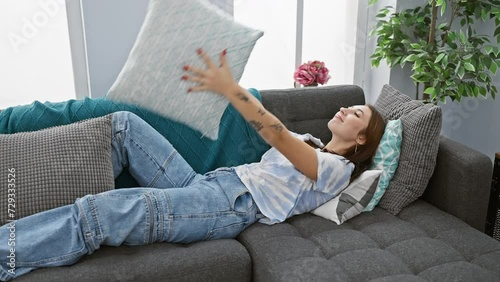 A relaxed woman lounging on a gray sofa tossing a turquoise pillow with a plant and sofa cushions in the background. photo
