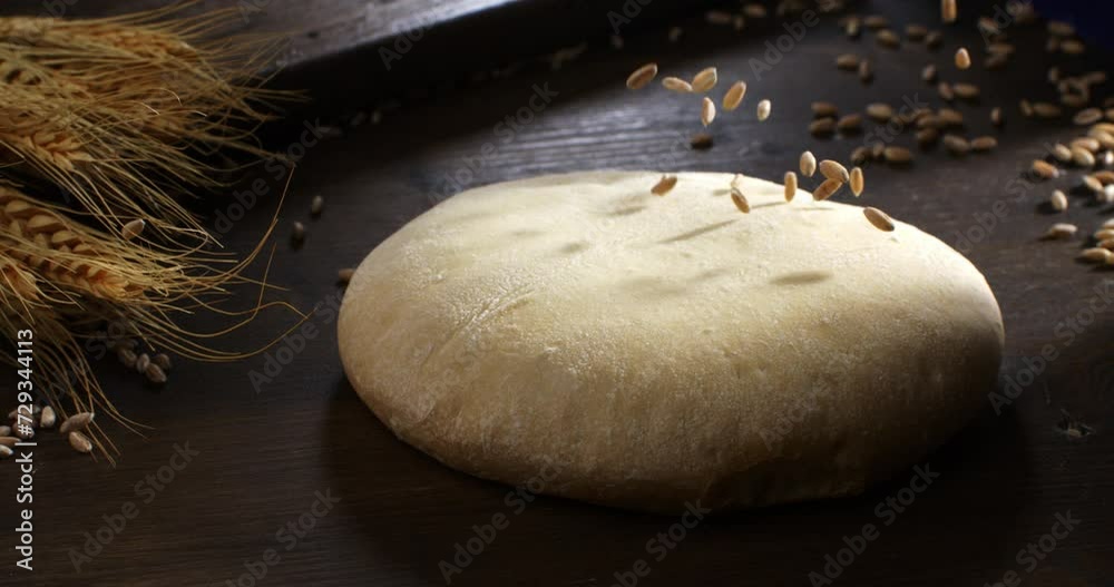 Super slow motion macro of wheat grain seeds are falling on raw loaf of dough while making homemade bread, pasta or pizza on rustic wooden table in traditional bakery kitchen.