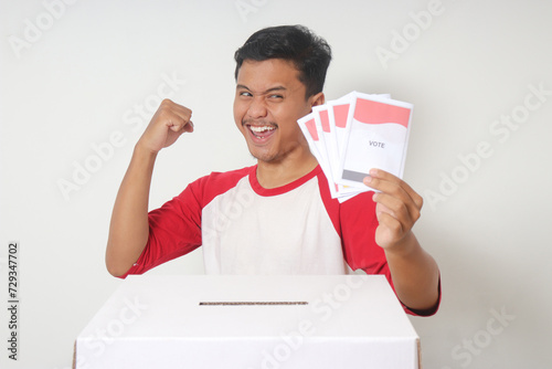 Portrait of excited Asian man inserting and putting the voting paper into the ballot box. General elections or Pemilu for the president and government of Indonesia. Isolated image on white background photo