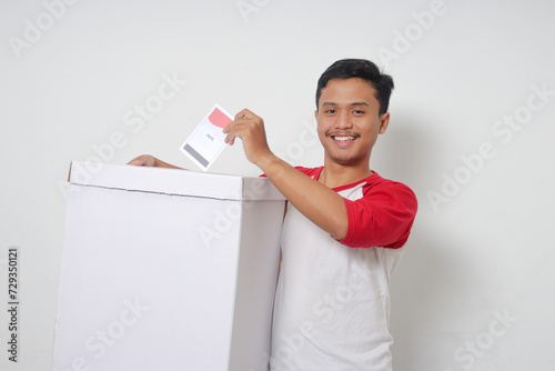 Portrait of excited Asian man inserting and putting the voting paper into the ballot box. General elections or Pemilu for the president and government of Indonesia. Isolated image on white background photo