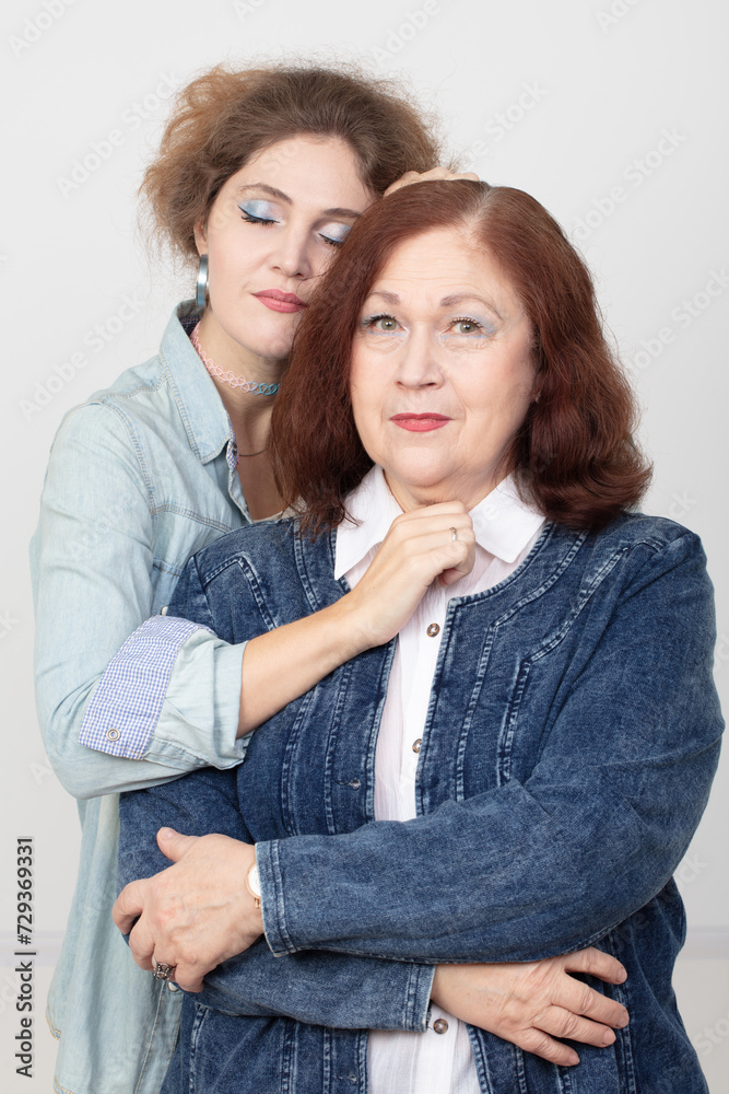 An elderly mother and an adult daughter are looking at the camera. The daughter-in-law gently hugs her mother-in-law. An elderly mother and an adult daughter are looking at the camera. The daughter-in-law gently hugs her mother-in-law.