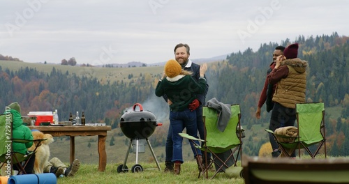 Group of travelers have fun during barbecue on hilltop. Happy multiethnic friends dance resting outdoors during vacation trip. Mountains landscape and forest in the background. Outdoor enthusiasts.