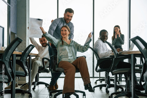 Fun at break time. Riding on a chair, smiling. Team of office workers are together indoors