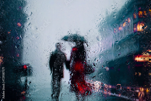 View through glass window with rain drops on blurred reflection silhouettesof a man and girl in walking on a rain under umbrellas and bokeh city lights, night street scene. Focus on raindrops on glass