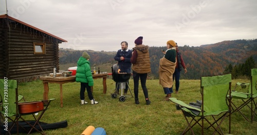 Multicultural family together cook food on grill and have fun on beautiful hill. Group of hiking buddies stopped to rest after long expedition at mountains. Concept of nature discovery and tourism.