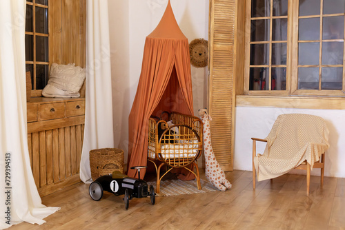 Rustic playroom interior with rattan crib with canopy, wicker baskets, toys and window. Wickered cradle bed with baldachin in newborn room in scandi style at home. Black racing car toy in a child room photo