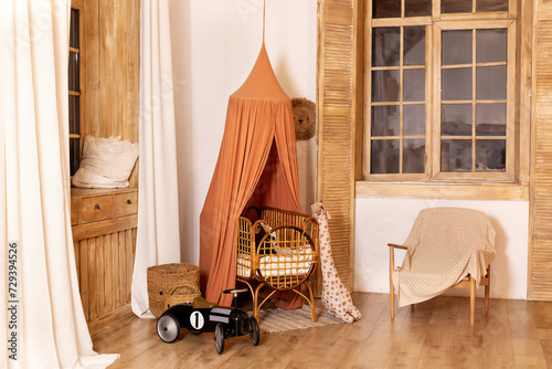 Rustic playroom interior with rattan crib with canopy, wicker baskets, toys and window. Wickered cradle bed with baldachin in newborn room in scandi style at home. Black racing car toy in a child room photo