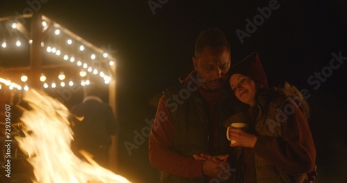 Multiethnic couple drinks tea, kisses and hugs while standing near the fire outdoors during vacation trip. In the background traveling friends rest in stylish wooden gazebo decorated with light bulbs.