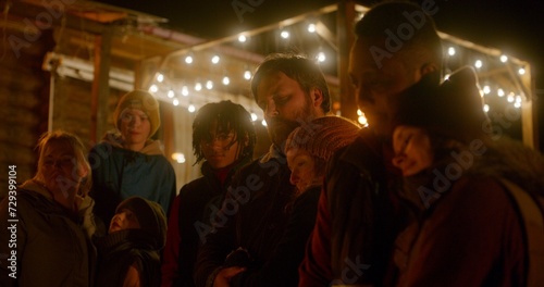 Group of multiethnic tourists talk while standing near the bonfire outside of wooden holiday cottage at night. Diverse friends or family resting outdoors during vacation in countryside or mountains.