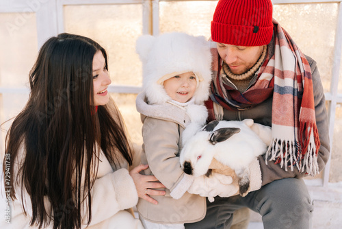 Front view of happy young family, father, mother and adorable son in warm clothes hugging white funny rabbit standing by house on sunny winter day. Concept of excursion to eco-farm, life in village