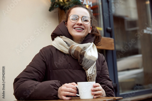 Portrait of young woman sitting outdoor, at terrace and drinking hot warming tea. Delight and smiling customer. Concept of medicine and beauty, health care and contemporary confidence.