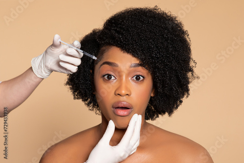 Surprised black woman getting facial injection, beige background
